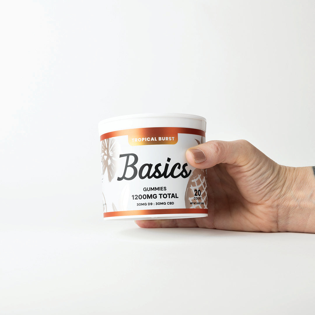 Hand holding a container labeled 'Basics' with orange and white design on a white background. 1200mg total cannabinoids. 30mg Nano D9 THC + 30mg CBD.
